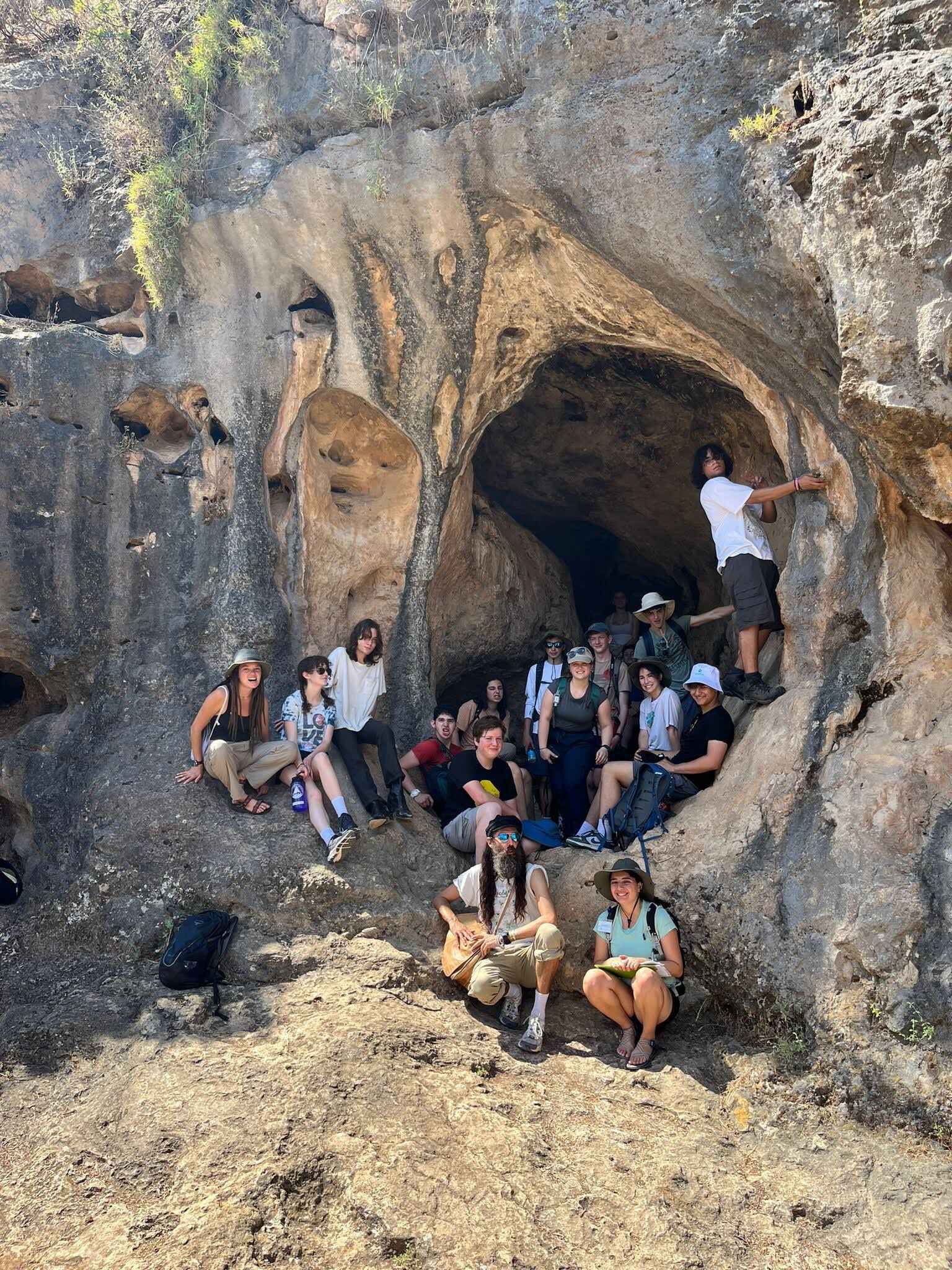 Group of individuals seated near a large rock cave, smiling outdoors.