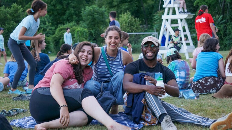 Group of three individuals sitting on grass, enjoying a summer day at a camp.