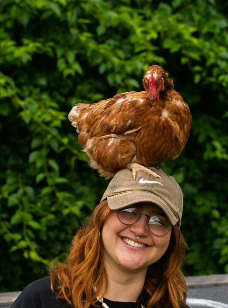 A person with glasses smiles, a chicken perched on their cap.