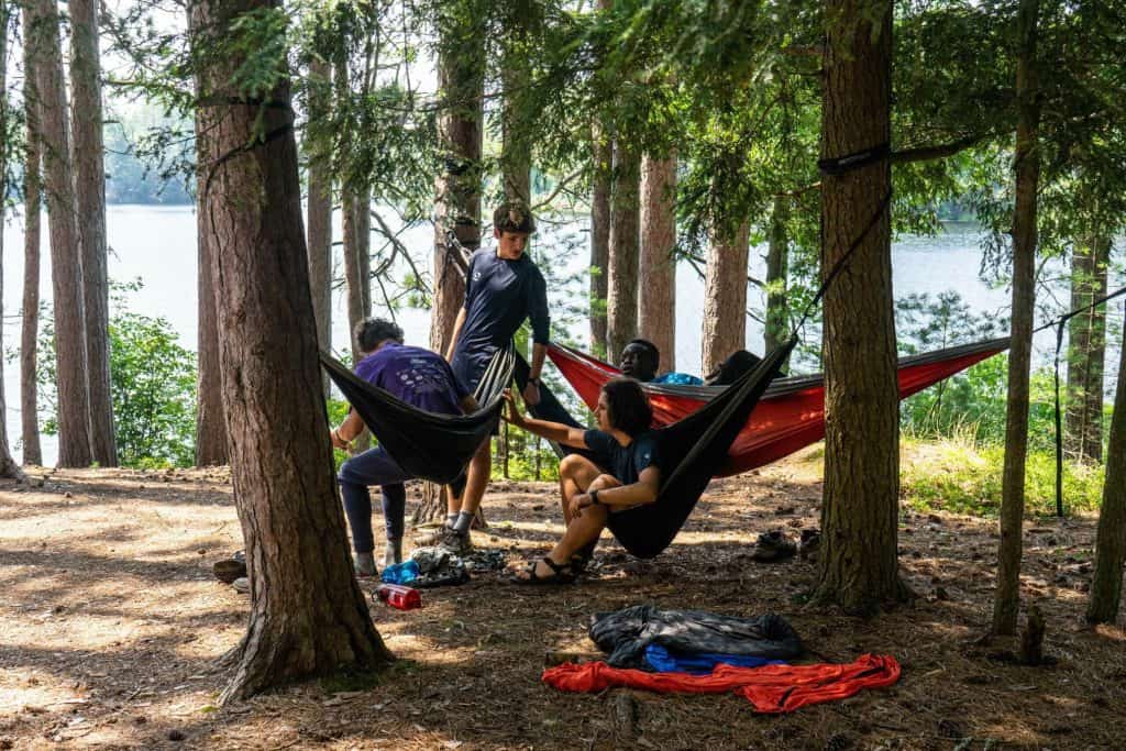 Young campers relaxing and socializing in hammocks at Eden Village Camp by a scenic lake surrounded by tall trees.