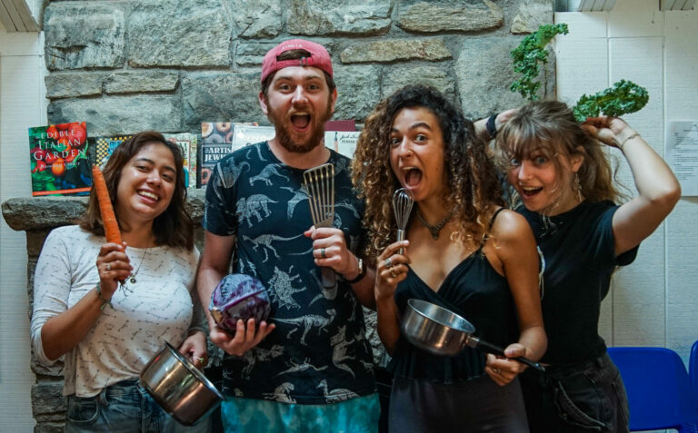 Four individuals gather in a kitchen, excitedly holding cooking utensils and ingredients.