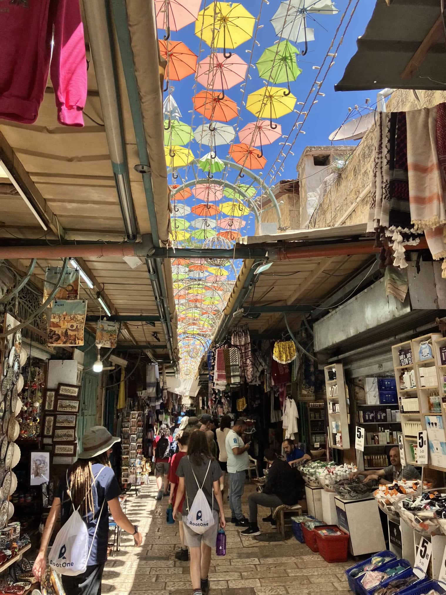 A lively market street with colorful umbrellas overhead and people browsing shops.