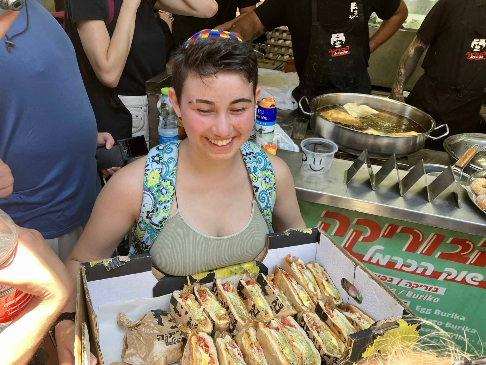Person smiling while holding a box of food at a bustling market.