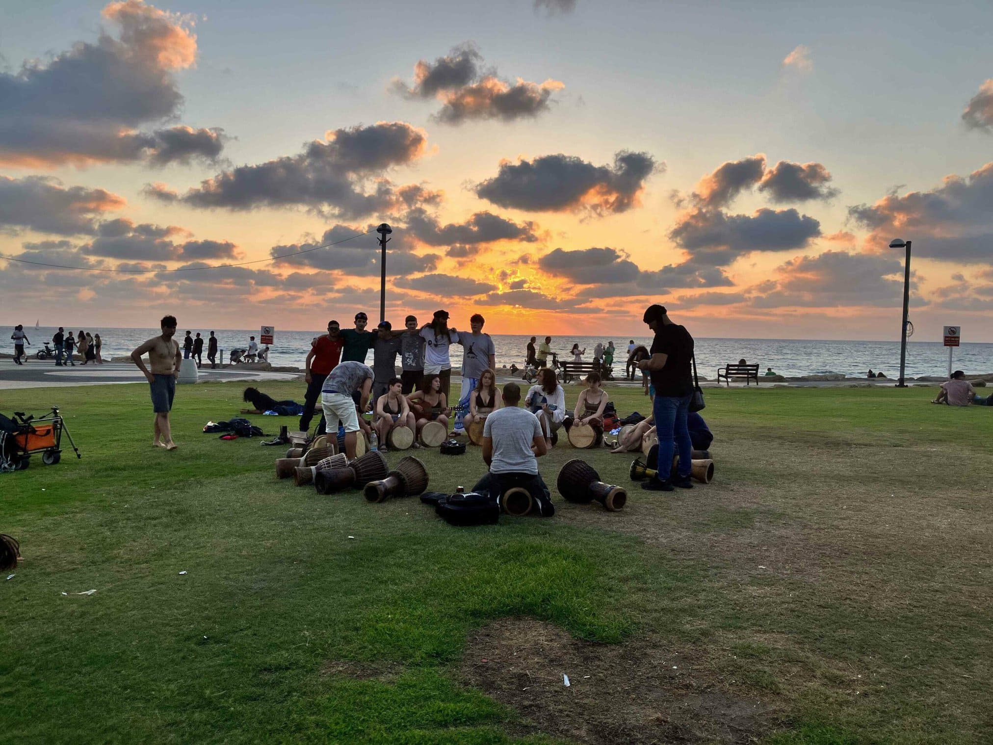 Group of people drumming on a beach at sunset, with onlookers nearby.