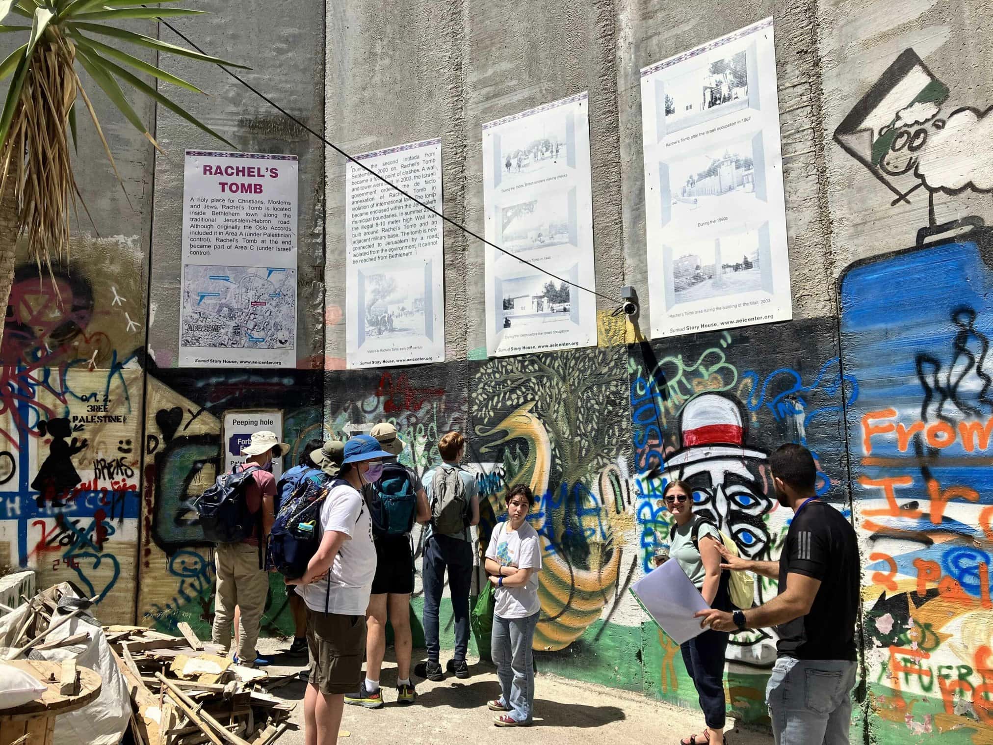 Group of people engaging with artwork and information about Rachel's Tomb on a graffiti-covered wall.