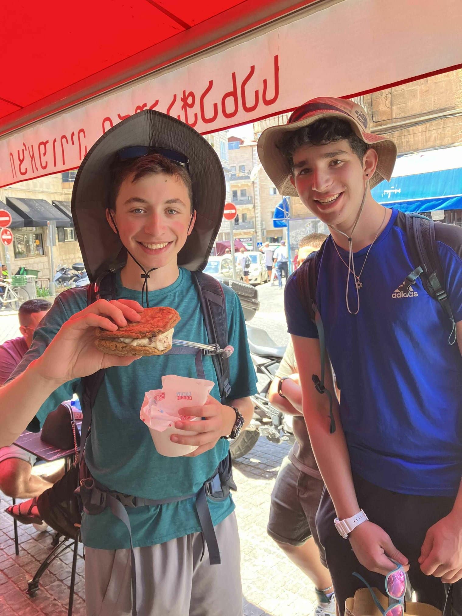 Two smiling individuals holding food items under a shaded awning in a busy market.