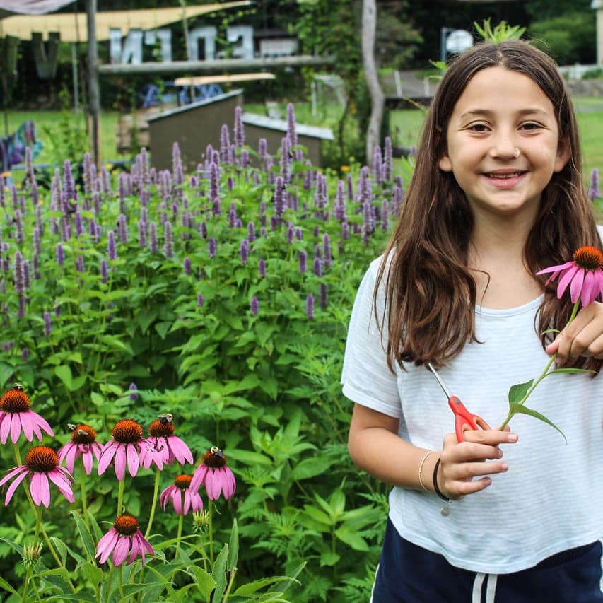 A smiling child holds a flower in a vibrant garden of purple and pink blooms.