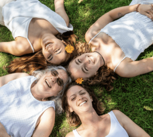 Four individuals lying on grass, smiling and enjoying a sunny day outdoors.