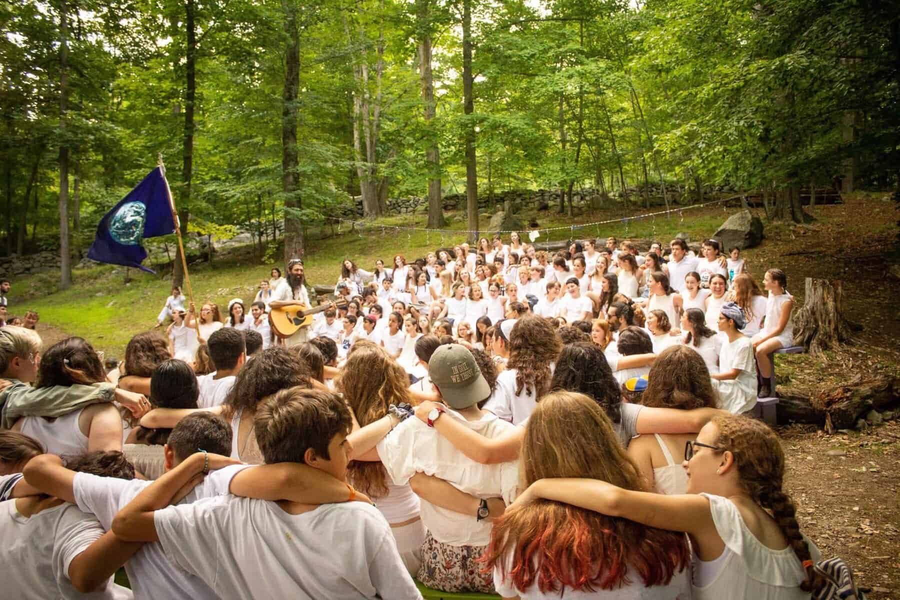 Vibrant outdoor camp gathering at Eden Village Camp with children singing and playing guitar surrounded by lush green trees in a forest setting.