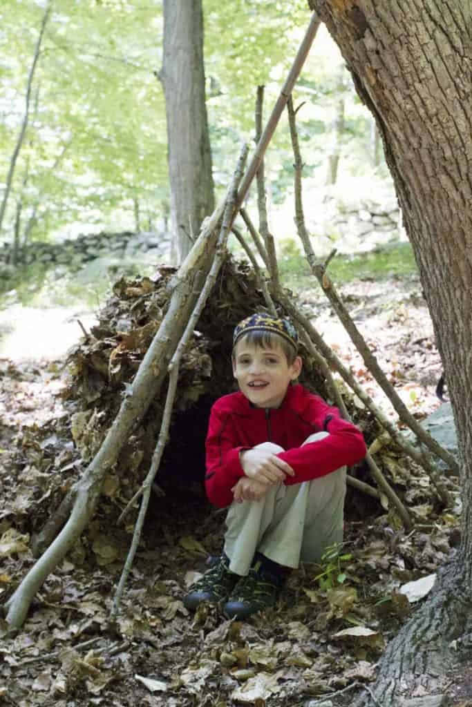 Handmade outdoor leafy shelter with a young boy sitting inside, surrounded by woodland trees and fallen leaves at Eden Village Camp.