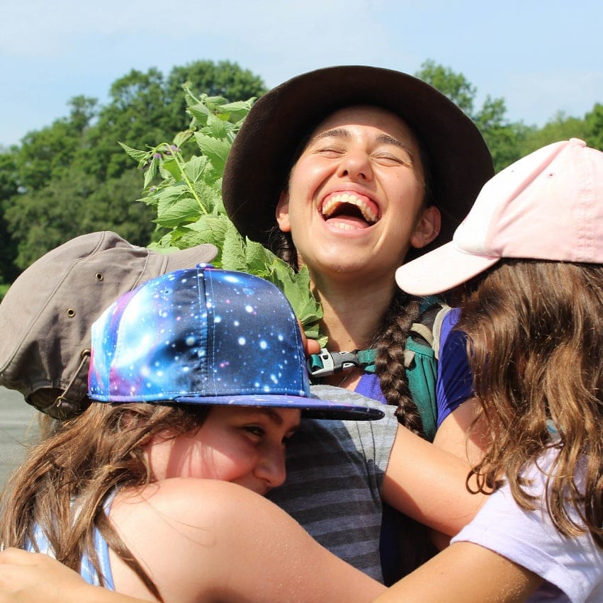 A happy individual wearing a wide-brimmed hat embraces participants outdoors.