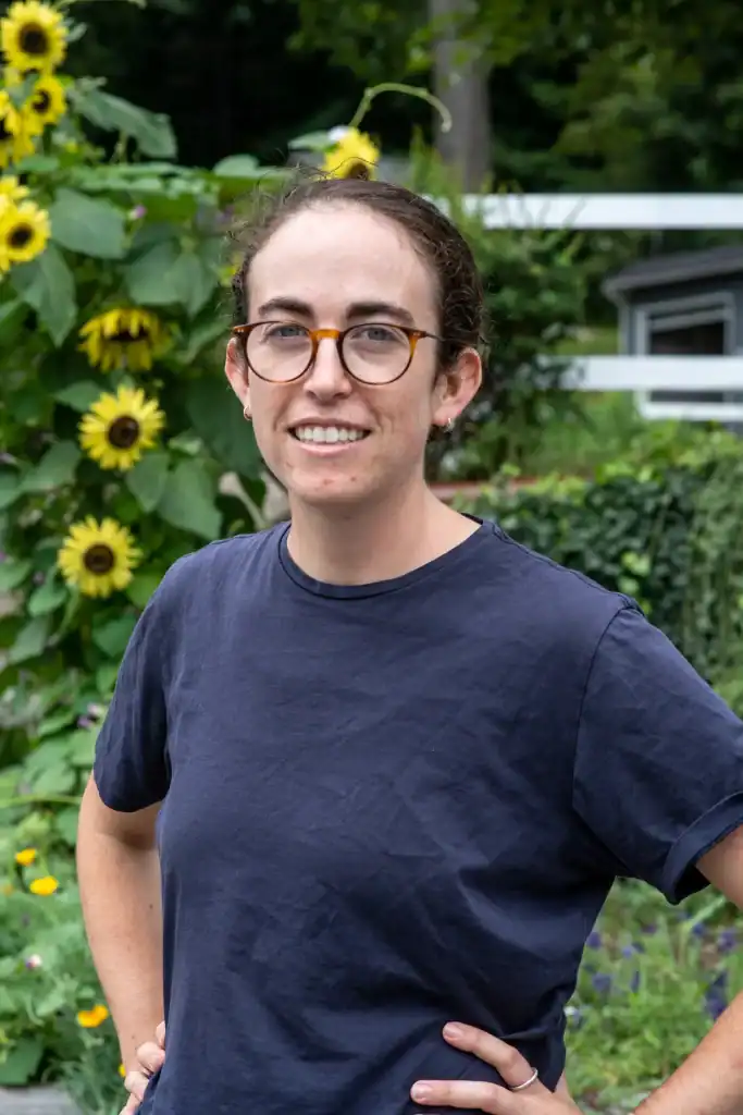 Youth camp counselor at Eden Village Camp, surrounded by vibrant sunflowers, smiling confidently in a natural outdoor setting, promoting eco-friendly and sustainable camping experiences.